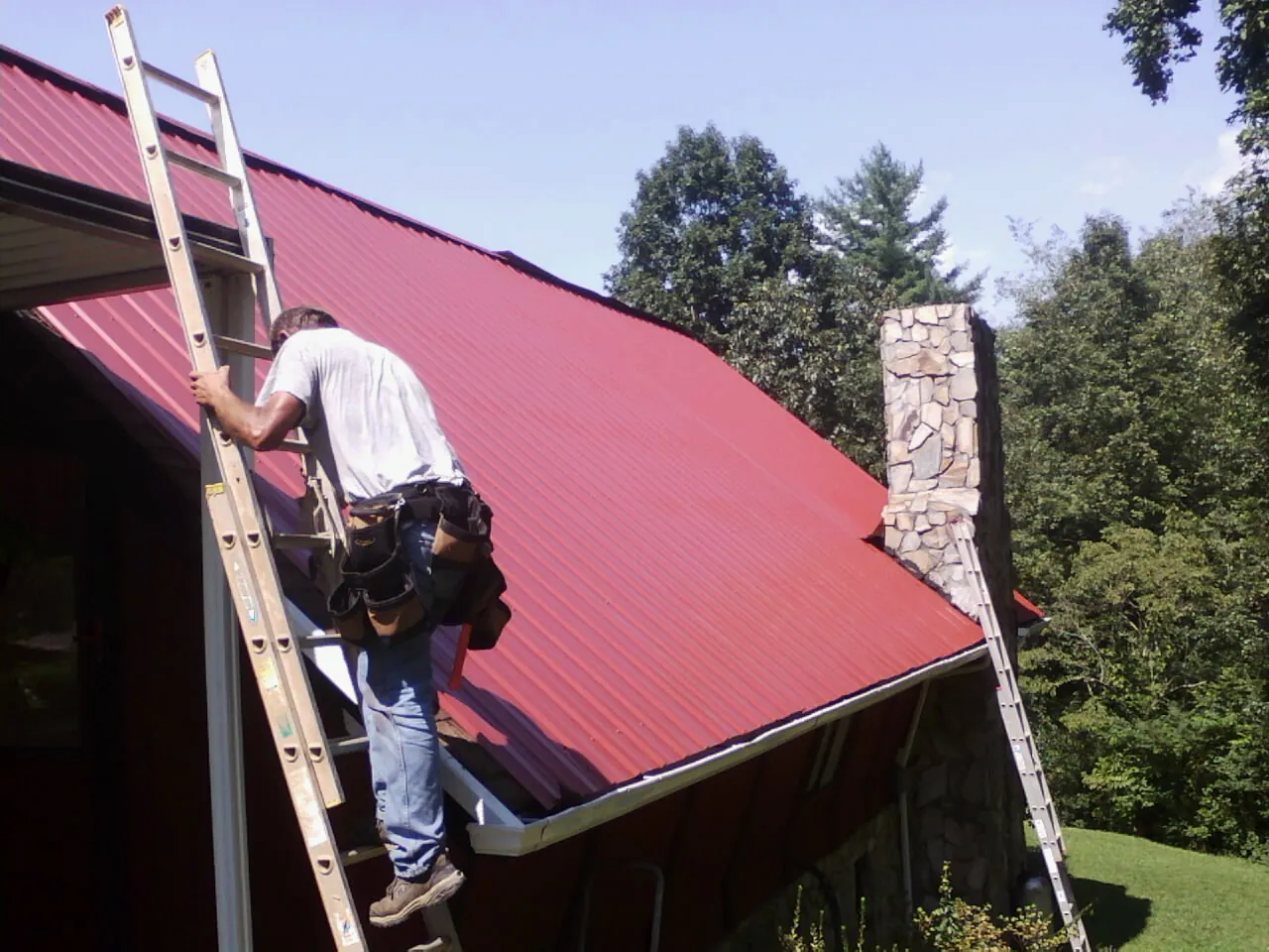 Roofer on a ladder beside a red metal roof and stone chimney — Soffit & Fascia Repair by Roofing Brothers in Latrobe